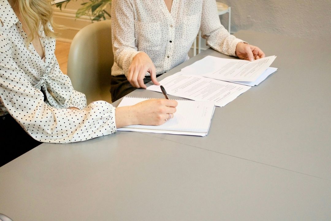 Woman signing on white printer paper beside woman about to touch the documents